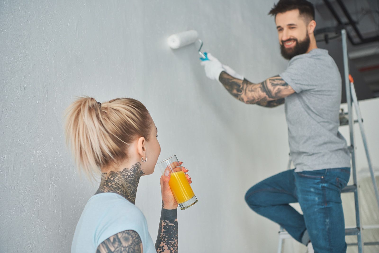 Young Woman Drinking Juice While Boyfriend On Ladder Is Painting Residential Wall Eggshell Texture